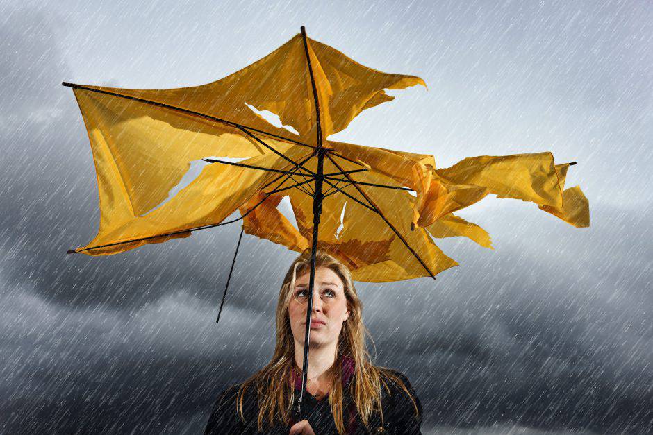 A beautiful young blonde woman sheltering under a torn and tattered yellow umbrella looks up miserably as she waits for a thunderstorm to pass.