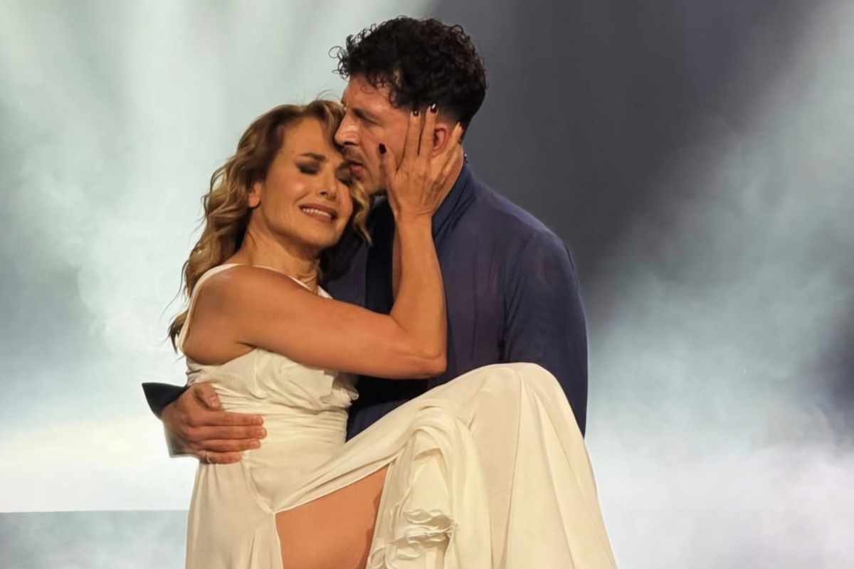 barbara d'urso con abito bianco e occhi chiuso in braccio a Pasquale la rocca con camicia che le bacia la fronte e sfondo fumoso