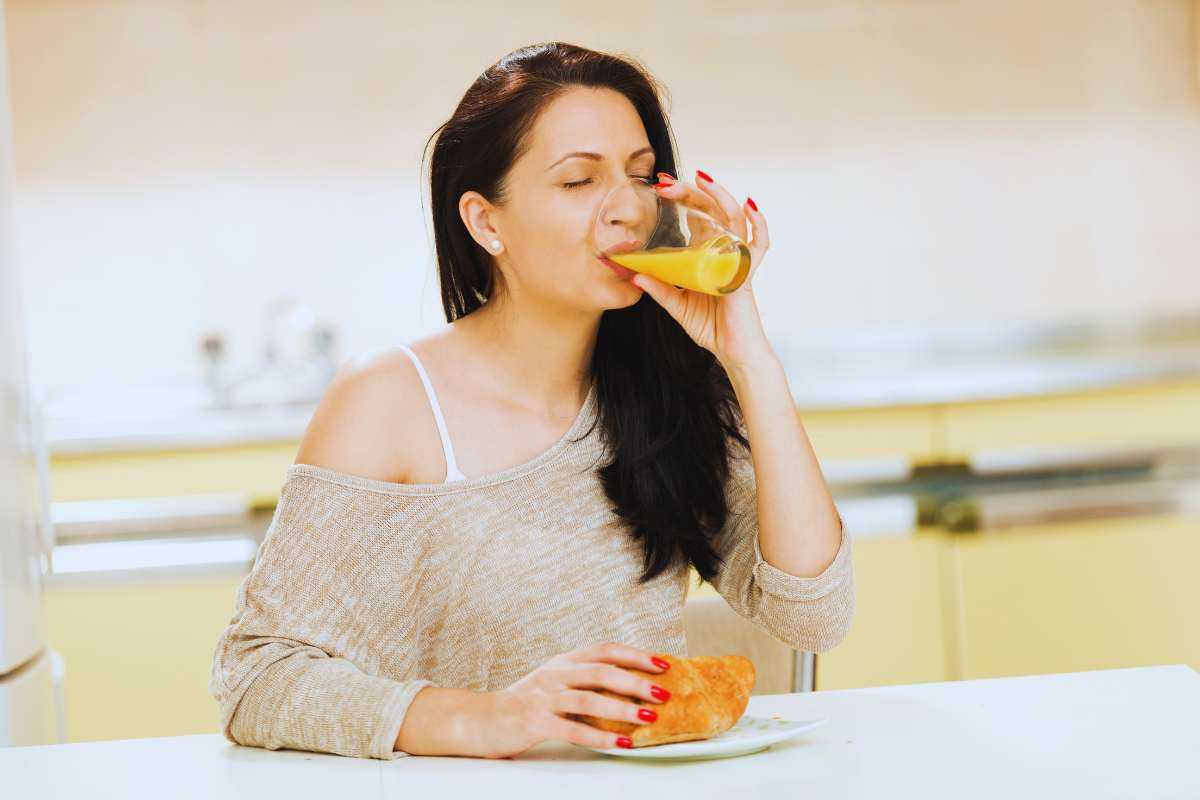 Donna che beve un succo di frutta seduta al tavolo con davanti a sé un cornetto