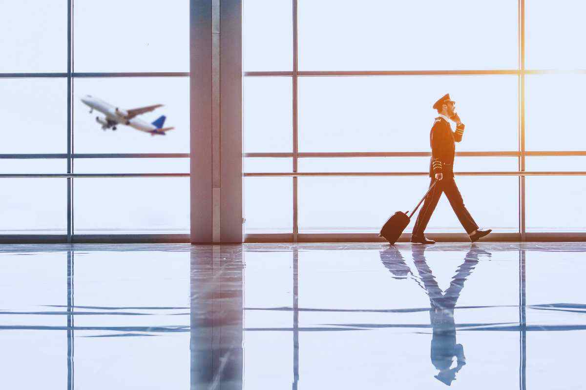 Membro dell'equipaggio di un aereo cammina in aeroporto con indosso la divisa parlando al cellulare, dietro di lui una vetrata con un aereo in volo