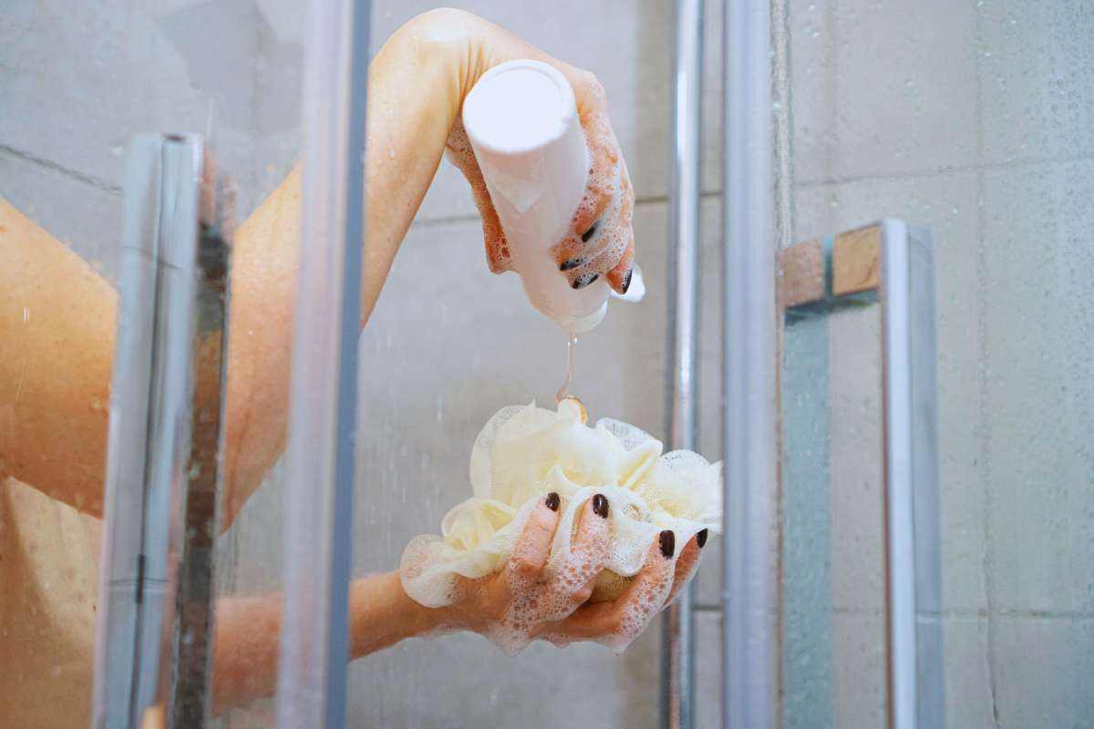 Una doccia aperta da cui escono le mani di una persona che tiene con una mano un barattolo di bagnoschiuma e con l'altra una spugna