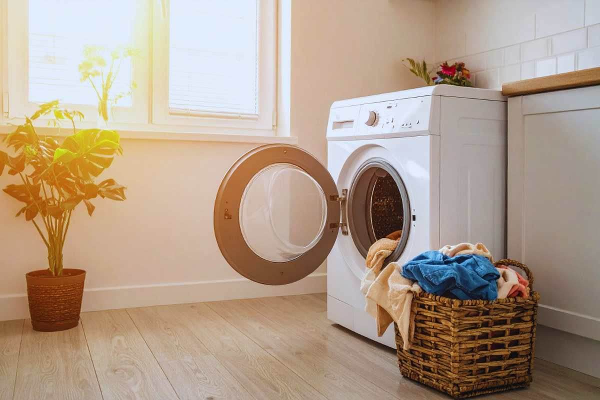 Una lavatrice con lo sportello aperto accanto dei panni da lavare, sullo sfondo una pianta e una finestra