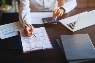 Uomo che scrive su un foglio di carta seduto ad una scrivania in mezzo ad altre scartoffie